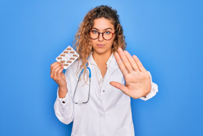 Young beautiful blonde doctor woman with blue eyes wearing stethoscope holding pills with open hand doing stop sign with serious