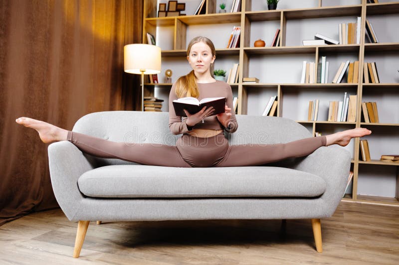 Young beautiful blonde woman in brown leggings and top sitting on a twine at home on the couch. The girl is reading a book sitting