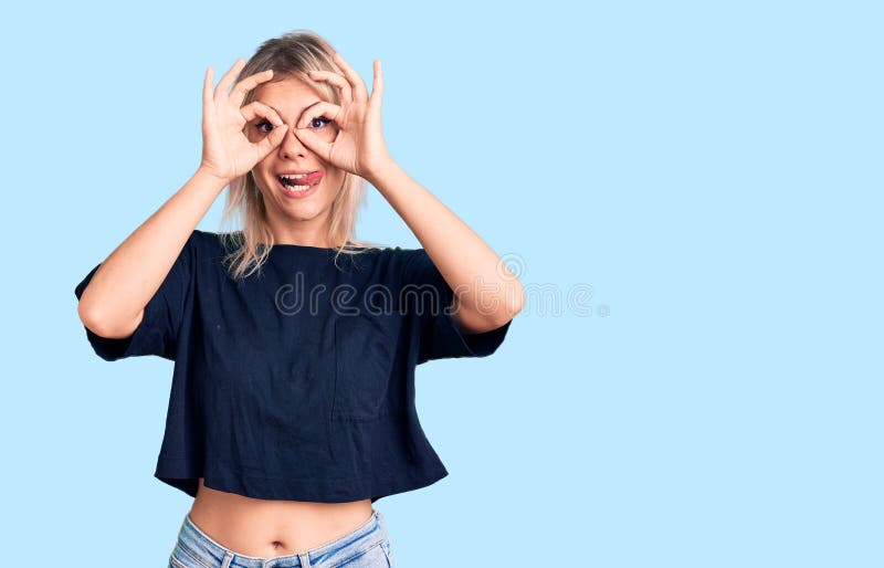 Young beautiful blonde woman wearing casual t-shirt doing ok gesture like binoculars sticking tongue out, eyes looking through