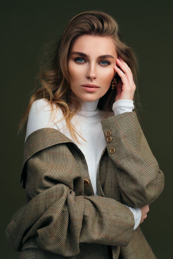A young beautiful blonde woman in a white turtleneck and a man`s jacket poses on a green olive background in the studio.