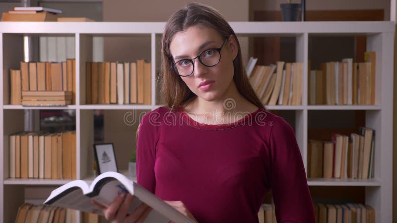 Young and beautiful brunette female student in eye glasses reading book watches into camera in library.