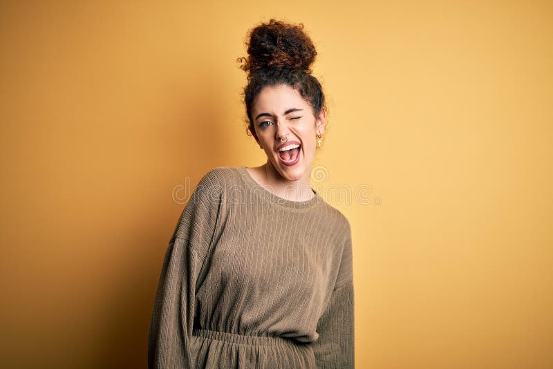 Young beautiful brunette woman with curly hair and piercing wearing casual dress winking looking at the camera with sexy
