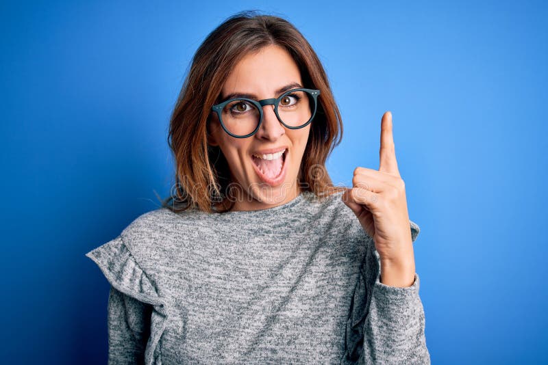 Young beautiful brunette woman wearing casual sweater and glasses over blue background pointing finger up with successful idea