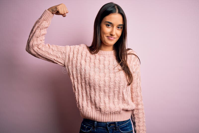 Young beautiful brunette woman wearing casual sweater over isolated pink background Strong person showing arm muscle, confident