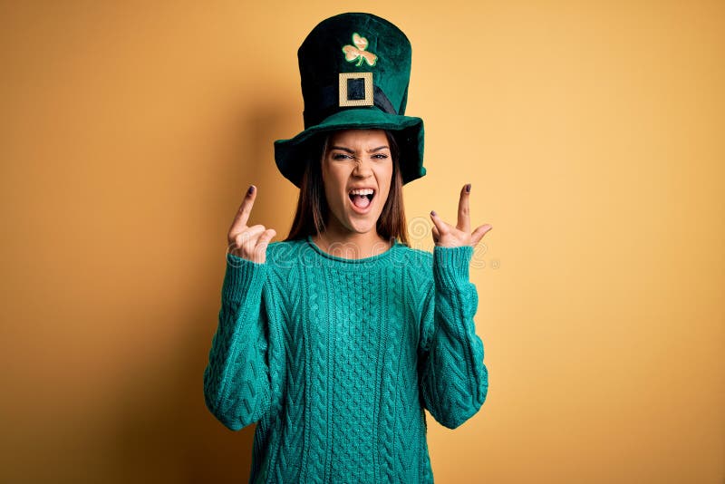 Young beautiful brunette woman wearing green hat with clover celebrating saint patricks day shouting with crazy expression doing