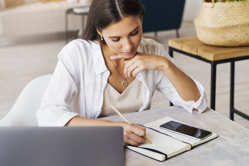 Young beautiful business woman making notes in notebook. Girl with dark hair in white shirt sits at table in front of laptop and writes with pencil. Distance education from home, re-profiling online. Young beautiful business woman making notes in notebook. Girl with dark hair in white shirt sits at table in front of laptop and writes with pencil. Distance education from home, re-profiling online.