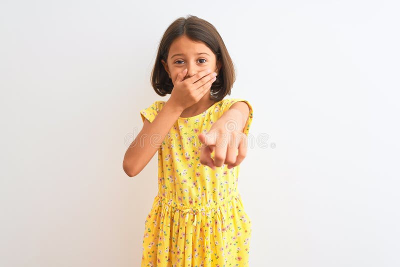 Young beautiful child girl wearing yellow floral dress standing over isolated white background laughing at you, pointing finger to