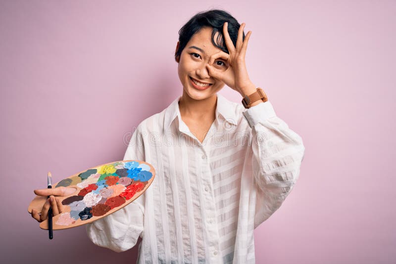 Young beautiful chinese artist woman painting using paintbrush and palette with colors with happy face smiling doing ok sign with