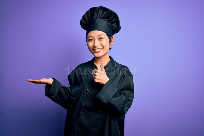 Young beautiful chinese chef woman wearing cooker uniform and hat over purple background Showing palm hand and doing ok gesture