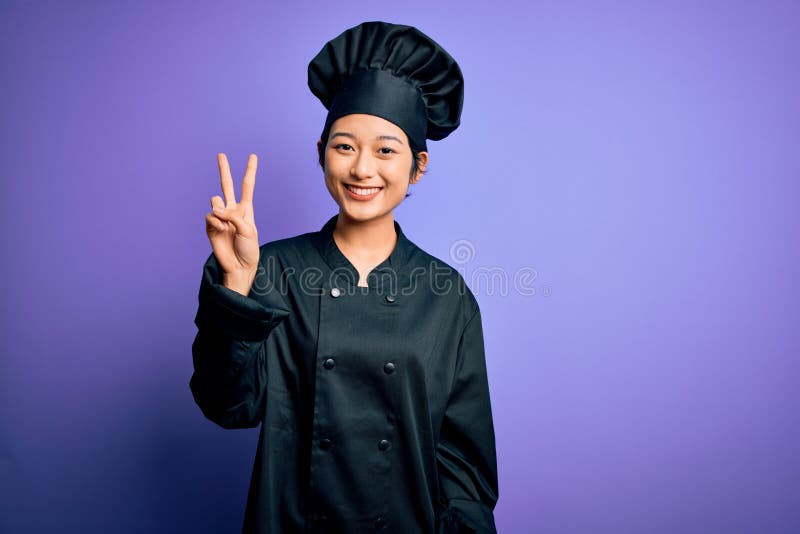 Young beautiful chinese chef woman wearing cooker uniform and hat over purple background smiling looking to the camera showing