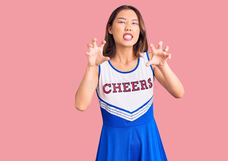 Young beautiful chinese girl wearing cheerleader uniform smiling funny doing claw gesture as cat, aggressive and sexy expression