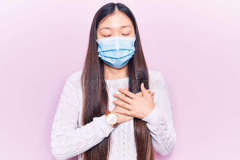 Young beautiful chinese woman wearing medical mask smiling with hands on chest, eyes closed with grateful gesture on face