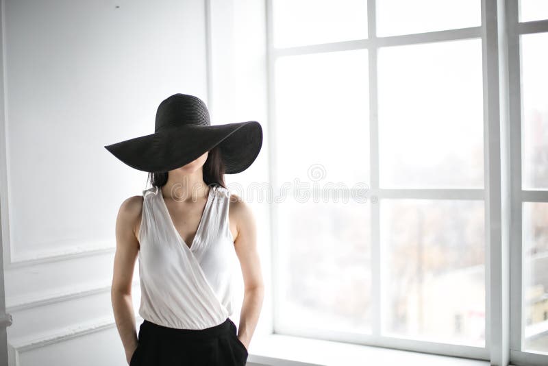 Young beautiful girl in the big black hat. Girl sits elegantly in a white chair.