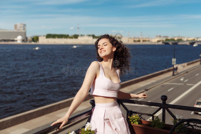 Young beautiful girl with curly hair in a pink dress is standing on the balcony and drinking coffee. Happy woman smiling and