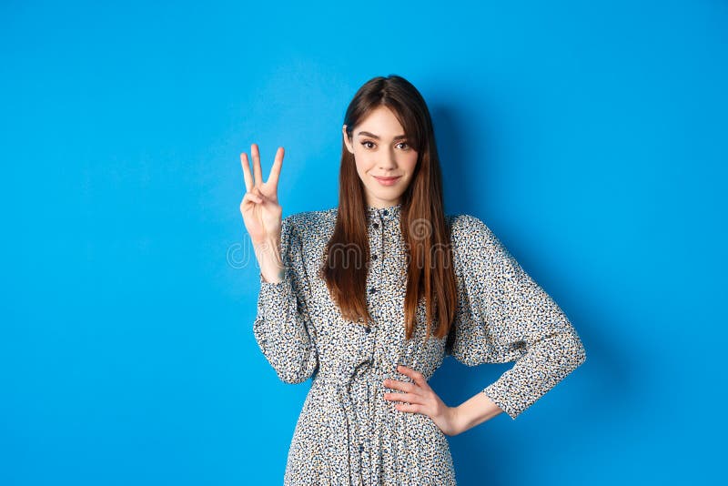 Young beautiful girl in dress with natural long hair, showing number three with fingers and smiling, standing against