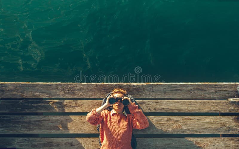 Young Beautiful Girl Lies On A Pier Near The Sea And Looks Through Binoculars On The Sky. Travel Search Journey Concept stock photography Young Beautiful Girl Lies On A Pier Near The Sea And Looks Through Binoculars On The Sky. Travel Search Journey Concept stock photography