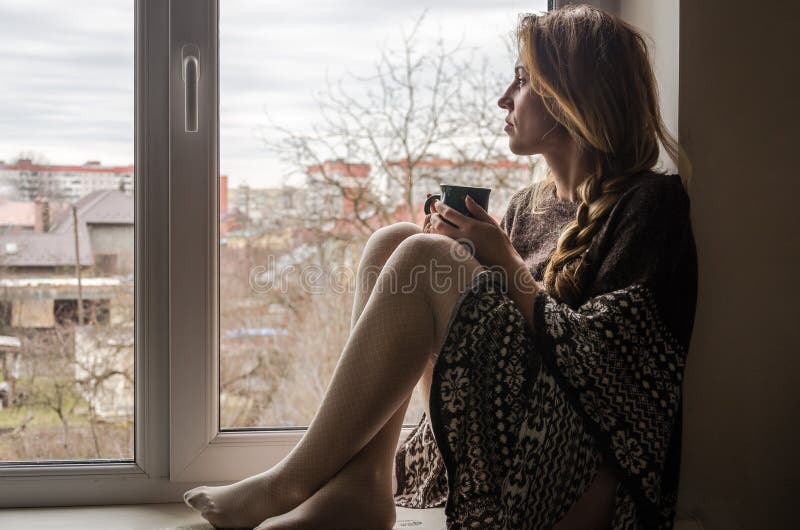 Young beautiful girl student sitting on a window sill at the window overlooking the city and drinking hot coffee from a mug