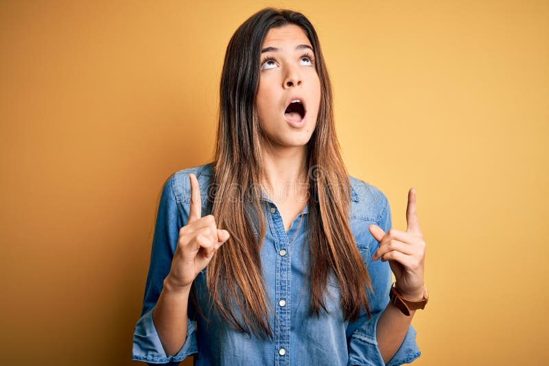 Young beautiful girl wearing casual denim shirt standing over isolated yellow background amazed and surprised looking up and