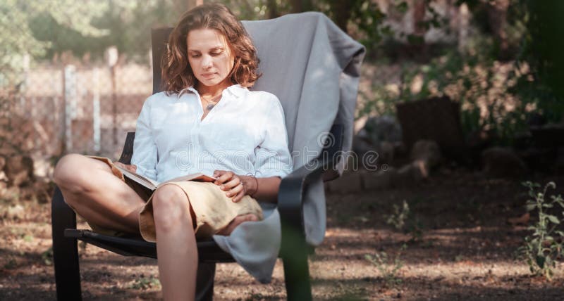 Young beautiful girl woman enjoys nature and reading a book in the summer in the garden of her house sitting on a chair