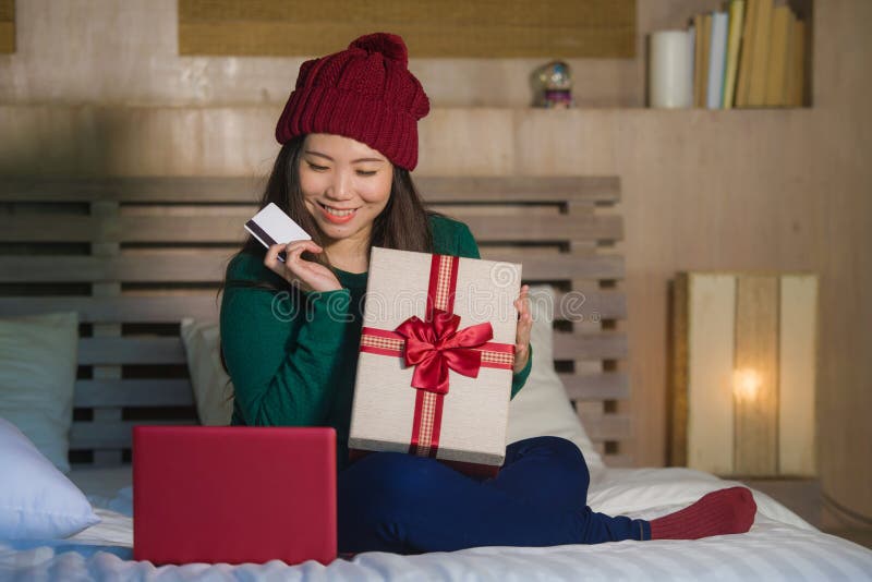 Young beautiful and happy Asian Chinese woman holding credit card and Christmas gift box shopping online xmas presents at home bed royalty free stock image Young beautiful and happy Asian Chinese woman holding credit card and Christmas gift box shopping online xmas presents at home bed royalty free stock image