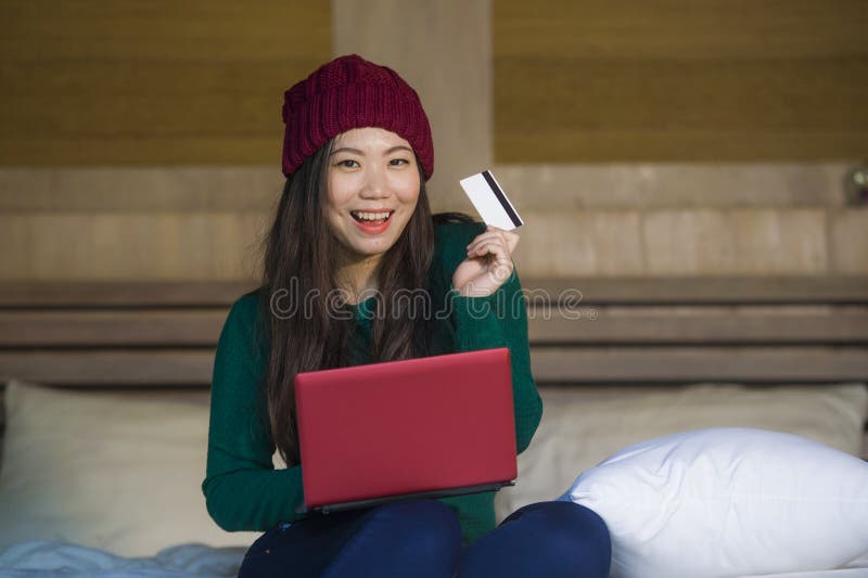 Young beautiful and happy Asian Chinese woman in winter hat sitting relaxed on bed holding credit card using laptop computer for i