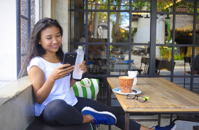 Young beautiful and happy Asian woman smiling relaxed enjoying breakfast sitting outdoors at coffee shop holding water bottle and mobile phone networking having social media internet fun. Young beautiful and happy Asian woman smiling relaxed enjoying breakfast sitting outdoors at coffee shop holding water bottle and mobile phone networking having social media internet fun