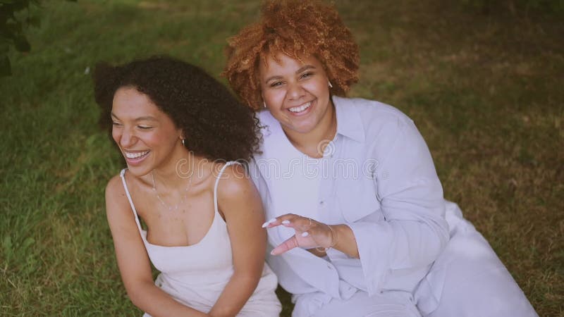 Young beautiful happy lesbian African American couple sitting on green grass hugging outside at nature summer park