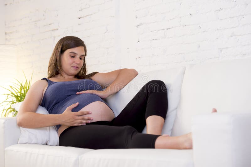 Young beautiful happy 8 or 9 months pregnant woman at home living room couch holding big belly