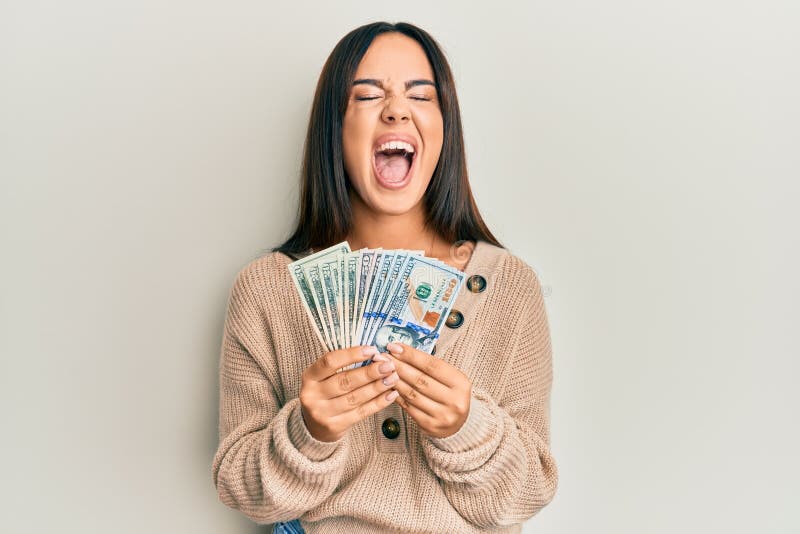 Young beautiful hispanic girl holding dollars angry and mad screaming frustrated and furious, shouting with anger looking up