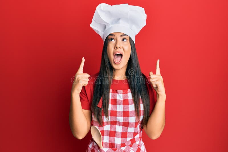 Young beautiful hispanic girl wearing baker uniform and cook hat amazed and surprised looking up and pointing with fingers and