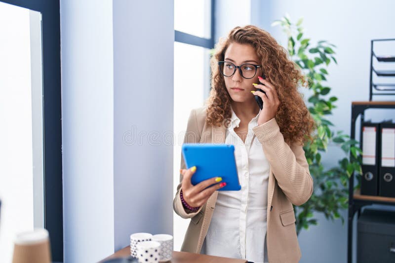 Young beautiful hispanic woman business worker using touchpad talking on smartphone at office stock photography