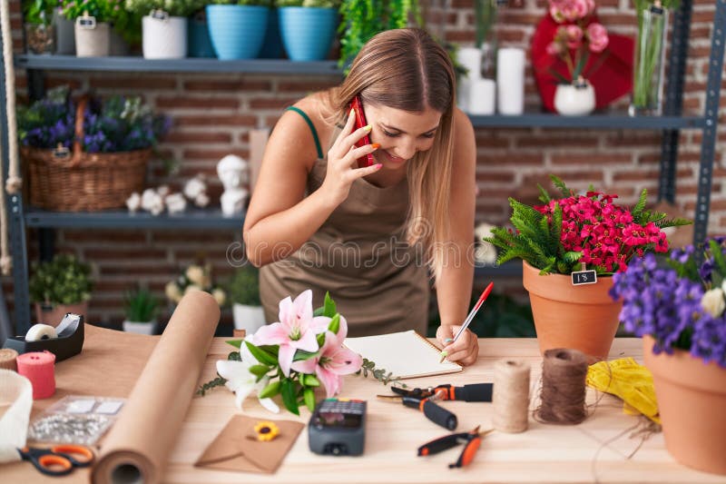 Young beautiful hispanic woman florist talking on smartphone writing on notebook at florist royalty free stock photography