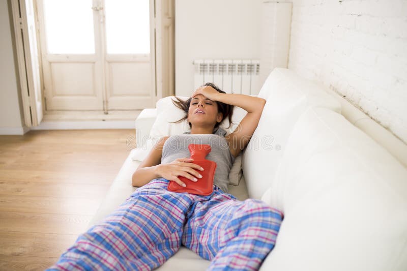 Young beautiful hispanic woman in painful expression holding hot water bottle against belly suffering menstrual period pain lying sad on home couch and having tummy cramp in female health concept. Young beautiful hispanic woman in painful expression holding hot water bottle against belly suffering menstrual period pain lying sad on home couch and having tummy cramp in female health concept