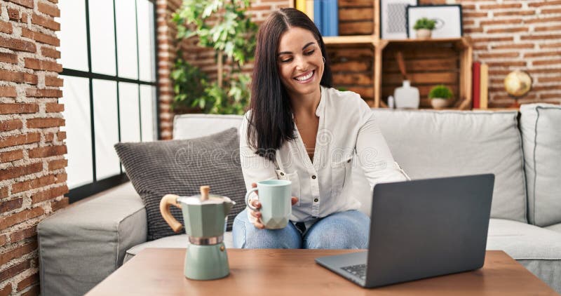 Young beautiful hispanic woman using laptop drinking coffee at home