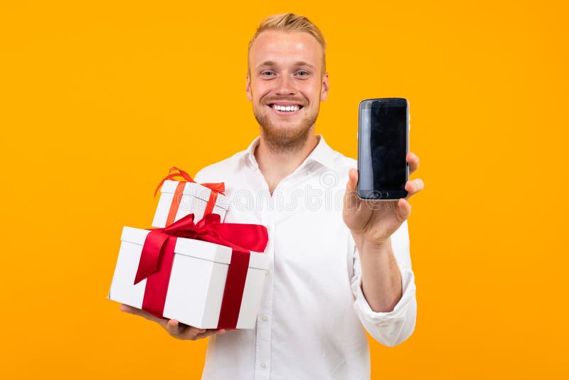 Young beautiful man with blonde hair in white shirt holds white box with gift and call the phone isolated on yellow