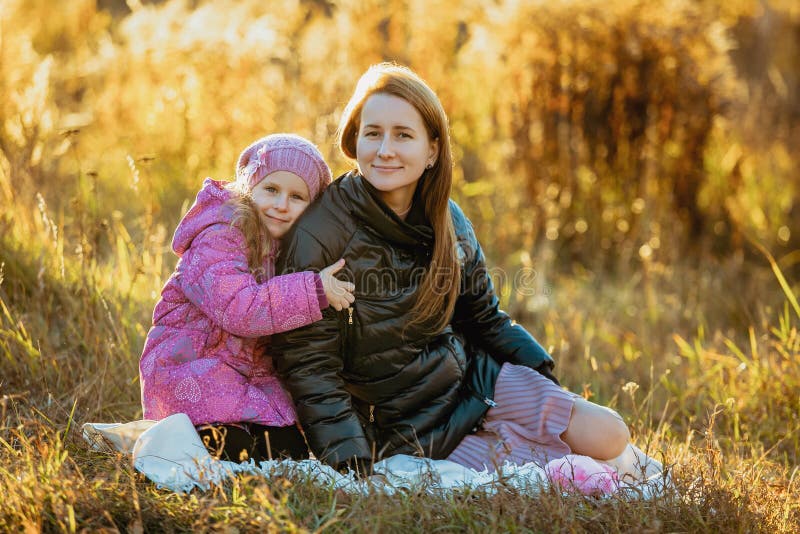 Young beautiful mother with her daughter on a walk on a sunny autumn day. They are sitting on a plaid on the grass, a daughter is stock photo Young beautiful mother with her daughter on a walk on a sunny autumn day. They are sitting on a plaid on the grass, a daughter is stock photo