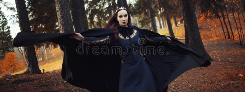 Young beautiful and mysterious woman in woods, in black cloak with hood, image of forest elf or witch