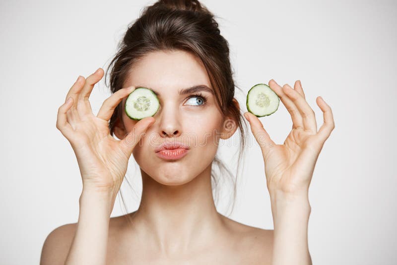 Young beautiful naked girl smiling hiding eye behind cucumber slice over white background. Beauty spa and cosmetology