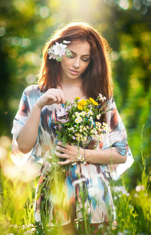 Young beautiful red hair woman holding a wild flowers bouquet in a sunny day. Portrait of attractive long hair female with flowers