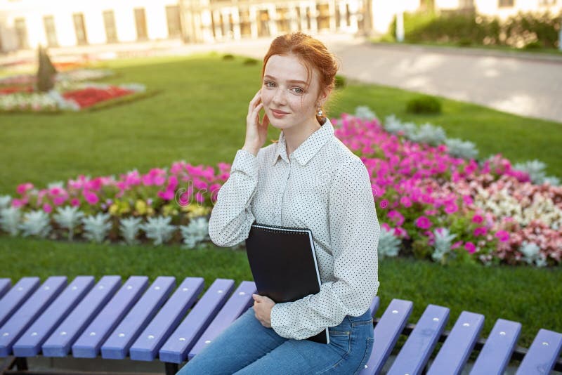 Young beautiful red-haired girl with freckles sitting on a bench near the university holds a notebook in her hands with homework. Portrait of a student. Back to school concept