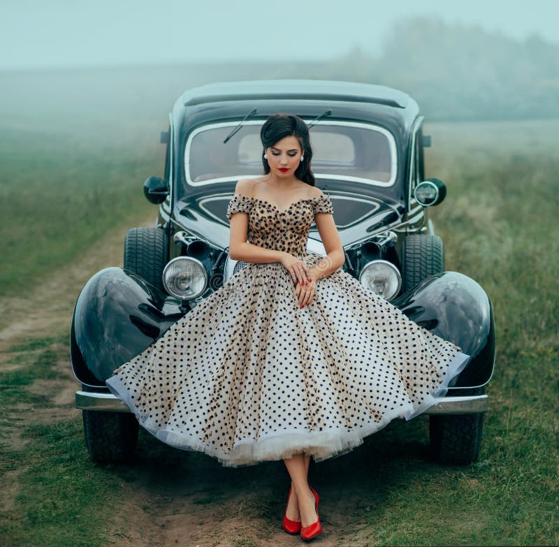 Young beautiful sexy woman in pin-up style clothes posing near black retro car. Polka dot white dress, vintage hairstyle. Red high heels. Background road green