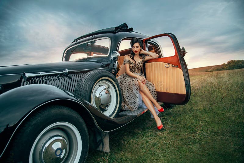 Young beautiful sexy woman in pin-up style clothes sits posing in black retro car. Polka dot white dress, vintage