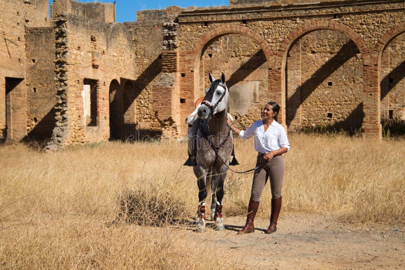 Young and beautiful Spanish woman with her thoroughbred horse. The woman is wearing a riding uniform and looks with love and