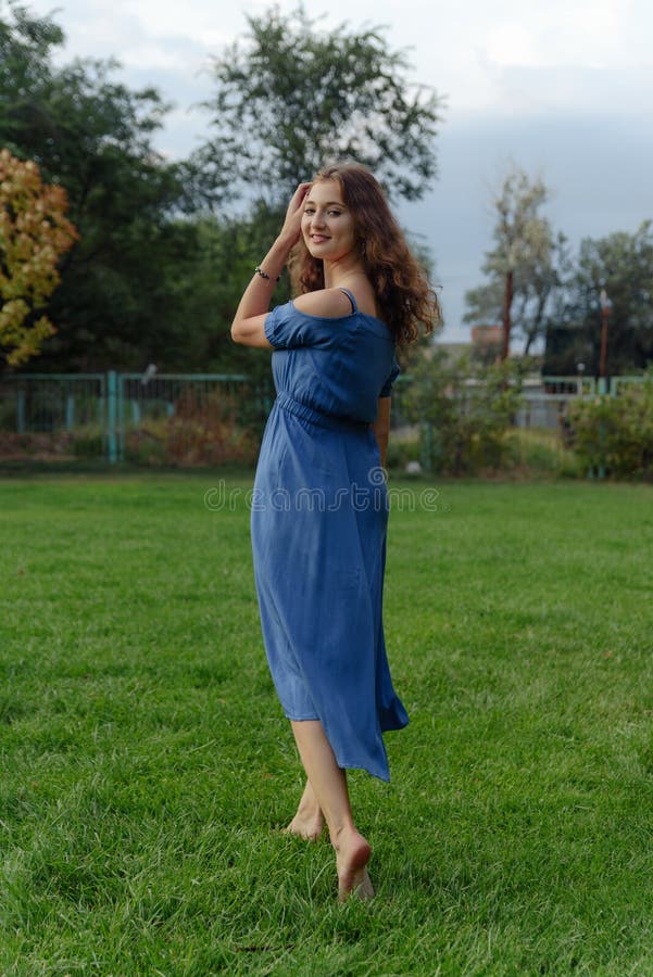 A Young Woman in a Blue Dress Walks Barefoot on the Grass in the Park ...