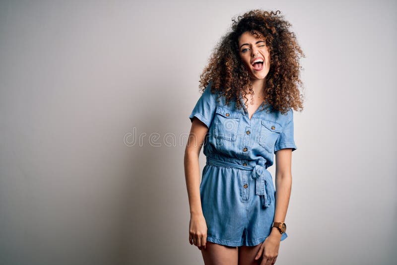 Young beautiful woman with curly hair and piercing wearing casual denim dress winking looking at the camera with sexy expression