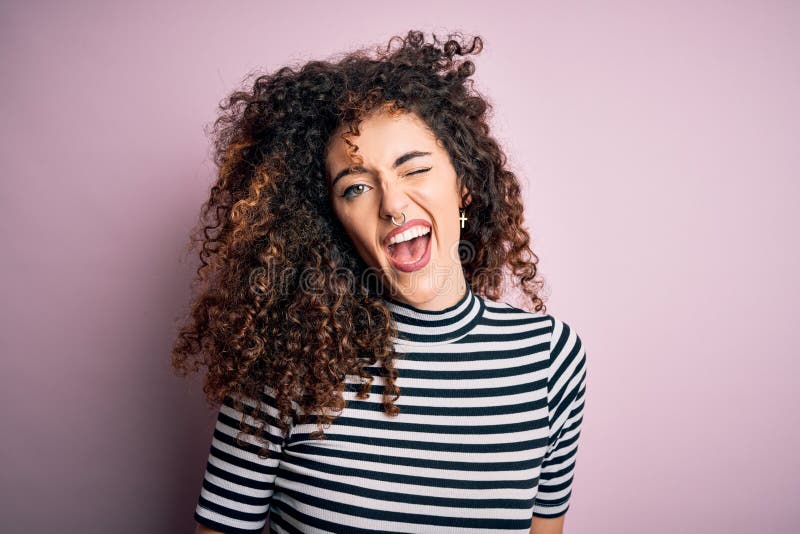 Young beautiful woman with curly hair and piercing wearing casual striped t-shirt winking looking at the camera with sexy