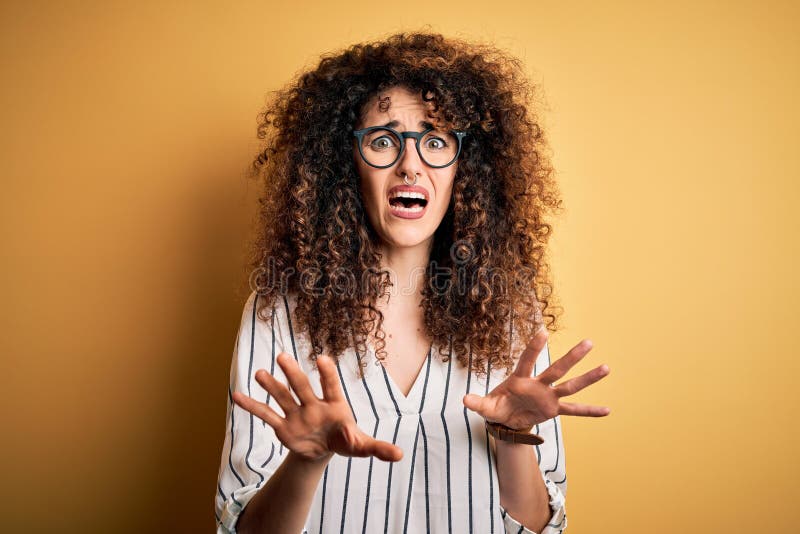 Young beautiful woman with curly hair and piercing wearing striped shirt and glasses afraid and terrified with fear expression