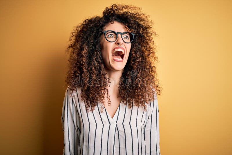 Young beautiful woman with curly hair and piercing wearing striped shirt and glasses angry and mad screaming frustrated and