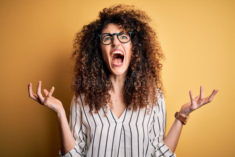 Young beautiful woman with curly hair and piercing wearing striped shirt and glasses crazy and mad shouting and yelling with