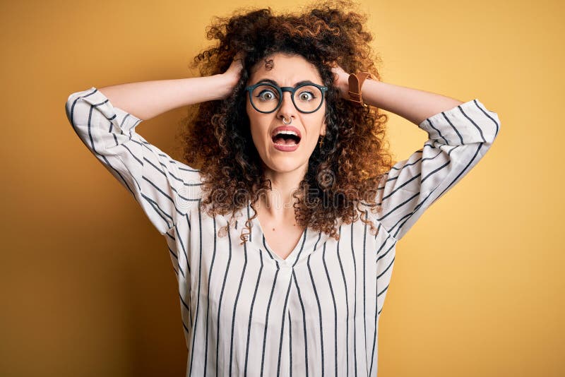 Young beautiful woman with curly hair and piercing wearing striped shirt and glasses Crazy and scared with hands on head, afraid